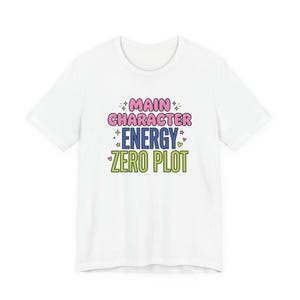 Puede incluir: Una camiseta blanca de manga corta con el texto MAIN CHARACTER ENERGY ZERO PLOT en letras de color rosa, azul y verde lima. El diseño incluye pequeños iconos decorativos de estrellas y corazones que rodean las palabras en un estampado.