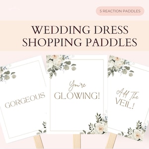 Może przedstawiać: Paddles do zakupów sukni ślubnych z napisami "Gorgeous", "You're Glowing!" i "Add The Veil!". Każdy paddle ma kwiatowe akcenty i jest na drewnianym patyku. Na górze obrazu widnieje napis "5 Reaction Paddles" i "Wedding Dress Shopping Paddles".