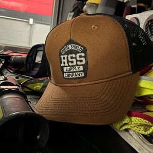 May include: Brown and black trucker hat with a mesh back. The front of the hat features a patch with the text "HOUSE SHIELD HSS SUPPLY COMPANY" in white and black.