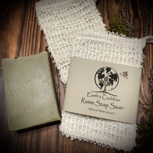 May include: A green bar of soap sits on a wooden surface next to a white, natural fiber pouch. The pouch is labeled "The Earth's Cauldron Ramie Soap Saver, natural fiber pouch".