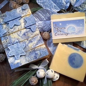 May include: Handmade soaps with a winter theme. One stack of soaps features blue snowflake designs, while others have a blue and white marbled pattern. One soap has a label that reads "A Storm is Coming". Decorative bells and ornaments complete the wintery scene.