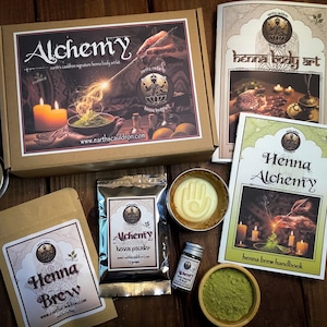May include: A collection of henna products, including a box labeled "Alchemy," a book titled "Henna Body Art," and a bag of "Henna Brew." Other items include a small jar, a tin with a hand design, and a bowl of green powder. The products are arranged on a wooden surface.