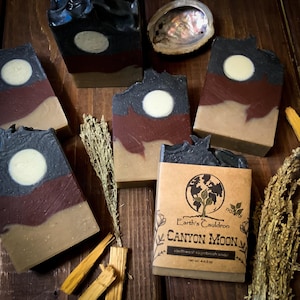 May include: Artisan soap bars with a layered design, featuring a dark gray top, a reddish-brown middle, and a tan base. Each bar has a white circular accent. One bar is labeled "Canyon Moon" with "southwest sagebrush soap".