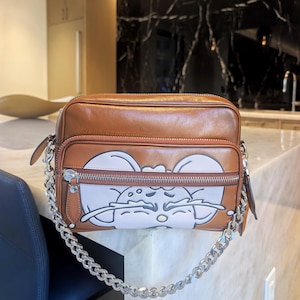 May include: A rectangular brown leather crossbody bag featuring a front zippered pocket with a light-colored graphic print of a stylized mouse face. The bag includes a silver-toned metal chain strap and matching silver hardware on the zippers.