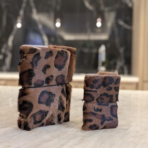 Può includere: Due polsini in pelle marrone e nera con stampa leopardata. I polsini presentano un motivo ripetuto di macchie marroni scure su uno sfondo marrone chiaro. Un polsino è più grande dell'altro.
