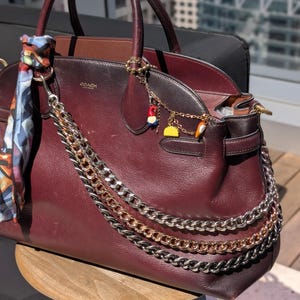 May include: A dark burgundy leather handbag featuring the Coach logo, adorned with a multi-colored patterned scarf tied to the handle, three layered silver and gold-tone metal chains, and a gold-tone charm bracelet with small colorful pendants.