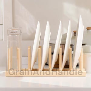 May include: A wooden dish rack with slots holding white plates. A clear glass is on the left. The text "GrainAndHandle" is printed on the base of the rack. The rack is made of light-colored wood and is on a white surface.