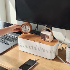 May include: A wooden-topped cable organizer with a white base, holding a small vintage radio and a wooden clock. A smartphone is charging in front of the organizer. The text "GrainAndHandle" is printed on the organizer.