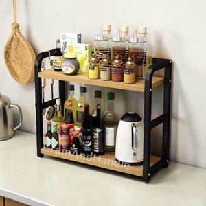 Puede incluir: Una estantería de cocina de dos niveles con estructura de metal negro y estantes de madera clara. Los estantes están llenos de varios frascos, botellas y una tetera eléctrica blanca. El texto "GrainAndHandle" es visible.