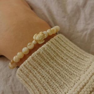 May include: A beaded bracelet featuring a turtle charm. The bracelet is made of cream-colored beads and a carved turtle accent. The bracelet is worn on a person's wrist, with a cream-colored sweater sleeve in the background.