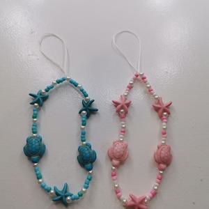 May include: Two beaded necklaces with a sea life theme. One necklace features turquoise turtles and starfish with blue and white beads. The other necklace has pink turtles and starfish with pink and white beads. Both have white string loops.