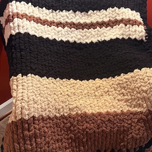 May include: A chunky knit blanket in a striped pattern. The blanket features alternating bands of black, cream, brown, and tan. The blanket is draped over a chair, showcasing its texture and color variations.