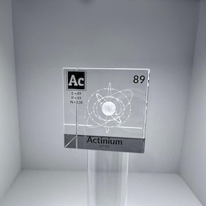 May include: A clear square glass block featuring a laser-etched atomic model of Actinium. The design includes the symbol Ac, atomic number 89, electron, proton, and neutron counts, and the name Actinium with the atomic mass 227.028 printed below.