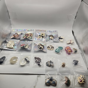 Pode incluir: Uma coleção de vários brincos, cada um embalado individualmente em pequenos sacos de plástico transparentes e seláveis. O sortido inclui diversas formas, cores e acabamentos metálicos, dispostos em filas sobre uma superfície branca.