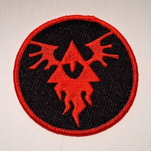 May include: Embroidered patch with a black background and a red emblem. The emblem is a stylized design with a central triangle and flame-like elements. The patch has a red border.