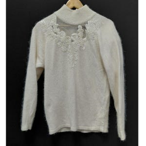 Può includere: Un maglione a maniche lunghe color crema con collo alto. La parte superiore del petto presenta un inserto in rete trasparente decorato con intricati ricami floreali bianchi e motivi a foglia che si estendono lungo lo scollo.