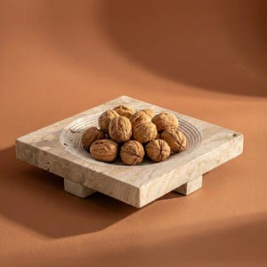 Könnte beinhalten: Eine quadratische, beige Marmorschale mit einer kreisförmigen Vertiefung, die mit Walnüssen gefüllt ist. Die Schale steht auf einem passenden quadratischen Sockel, mit einem warmbraunen Hintergrund. Die Schale ist ein dekoratives Stück für die Küche oder das Esszimmer.