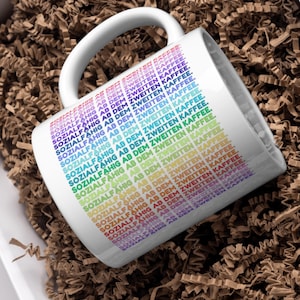 Könnte beinhalten: Weiße Keramik-Tasse mit gebogenem Henkel, mit dem Text "SOZIALFÄHIG AB DEM ZWEITEN KAFFEE" in einem Regenbogen-Farbverlauf. Die Tasse liegt in braunem Krepppapier in einer weißen Schachtel.