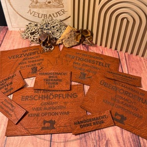Könnte beinhalten: Eine Sammlung brauner Lederetiketten mit eingraviertem Text und einer Nähmaschinen-Grafik. Die Etiketten zeigen Sätze wie "Handgemacht mit Koffein & Chaos" und "Gemacht aus purer Verzweiflung".