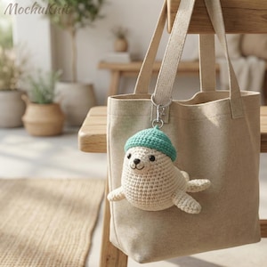 Op de afbeelding: Een beige draagtas met een gehaakte zeehond sleutelhanger. De zeehond is gebroken wit met een turquoise muts. De sleutelhanger is bevestigd aan de handgreep van de tas. Het Mochuknit-logo staat linksboven.