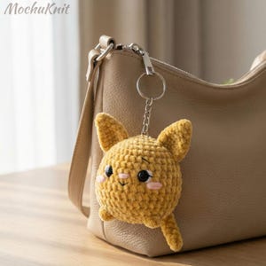 Könnte beinhalten: Ein handgefertigter gelber Häkel-Schlüsselanhänger in Form eines lächelnden Hasen mit schwarzen Augen und rosa Wangen. Der Schlüsselanhänger ist an einem silbernen Schlüsselring befestigt und hängt neben einer beigen Handtasche. Der Text "MochuKnit" befindet sich oben links.