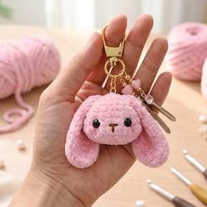 May include: A pink crocheted bunny keychain with black eyes and a brown nose. The keychain has a gold-colored clasp and decorative charms. The bunny is made of soft, textured yarn. The keychain is held in a hand.