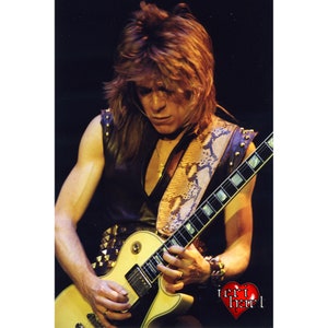May include: A musician playing a cream-colored electric guitar. The guitar has a black pickguard and silver hardware. The musician wears a black leather vest with studded accents and a snakeskin guitar strap. The image has a heart-shaped logo in the bottom right corner.
