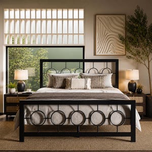 May include: A black metal bed frame with a headboard featuring circular and arched designs. The bed is made up with neutral-toned bedding and decorative pillows. Two nightstands with lamps flank the bed. The room has a large window and a neutral color palette.