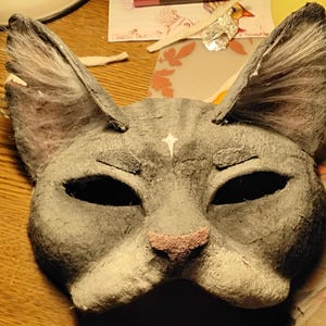 May include: A gray cat mask with large ears, black eye holes, and a pink nose. A white star is painted on the forehead. The mask is made of a textured material, possibly paper or cardboard, and is designed to be worn.