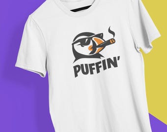 May include: White t-shirt with a graphic of a puffin wearing sunglasses and smoking a cigar. The word "PUFFIN'" is printed below the graphic in a bold, black font. The shirt is hanging on a wooden hanger.