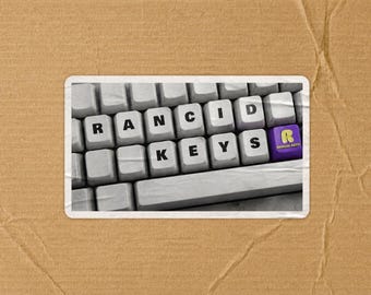 May include: A rectangular sticker featuring a black and white photograph of a computer keyboard. The keys are printed with the letters R, A, N, C, I, D, K, E, Y, S. A single purple key on the right side features a yellow letter R and the text RANCID KEYS.
