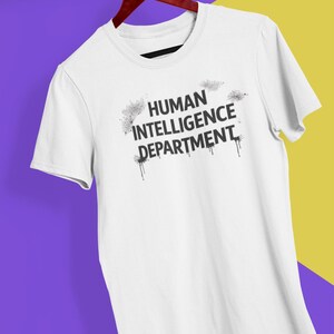May include: White t-shirt with the text "HUMAN INTELLIGENCE DEPARTMENT" in black, with a distressed, spray-paint style design. The shirt is hanging on a wooden hanger against a purple and yellow background.