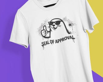May include: White t-shirt featuring a cartoon seal wearing sunglasses and giving a thumbs-up. The phrase "SEAL OF APPROVAL" is printed below the seal in a graffiti-style font. The shirt is hanging on a wooden hanger.