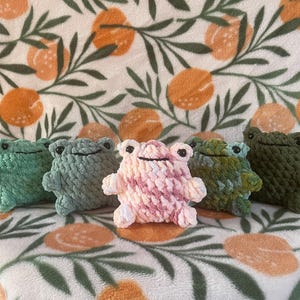 May include: Five handmade crochet frog plushies in shades of green, blue, and pink. The frogs are arranged on a patterned blanket with orange fruit and green leaves. Each frog has a smiling face.