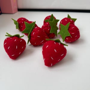 Puede incluir: Fresas hechas a mano de fieltro rojo con semillas cosidas en blanco y hojas de fieltro verde. Estas fresas decorativas están dispuestas sobre una superficie blanca.