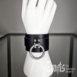 Könnte beinhalten: Ein breites Armband aus schwarzem Leder mit einem großen, polierten silberfarbenen Metallring, der durch zwei kleinere silberfarbene Metallschlaufen in der Mitte befestigt ist. Der Text "Crafts by nanna mary" ist unten rechts zu sehen.