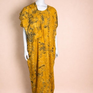May include: A mustard yellow, floor-length dress with a tie-dye pattern in shades of black and gray. The dress features a unique asymmetrical design with a draped, layered effect on the bodice. The dress is displayed on a white mannequin.