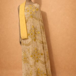 May include: A cream-colored formal gown with intricate gold embroidery and embellishments. A matching yellow scarf is draped over the shoulder. The dress is displayed on a mannequin against a neutral background.