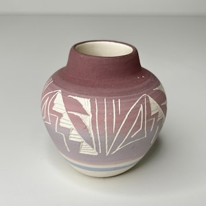 May include: A small, round ceramic vase with a wide body and a narrow neck. The vase is primarily a gradient of purple and mauve, with white geometric designs carved into the surface. A thin blue stripe encircles the base.