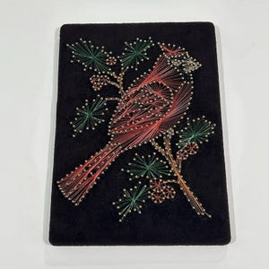 May include: A rectangular black velvet board featuring string art of a red cardinal bird perched on a branch. The design is created with silver nails and thin red, green, and brown thread, depicting the bird among pine needles and berries.
