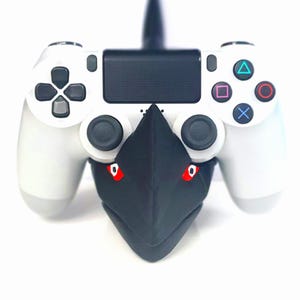 Può includere: Controller PlayStation 4 bianco con un accessorio nero a forma di uccello. L'accessorio ha occhi rossi e un becco. Il controller presenta il layout standard dei pulsanti.