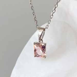 May include: A silver necklace featuring a rectangular, light pink gemstone pendant. The pendant is set in a silver frame and hangs from a delicate silver chain. The gemstone has a subtle sparkle, and the necklace is displayed against a white background.
