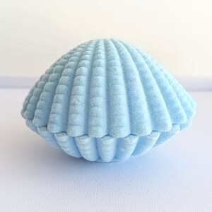 May include: A light blue, seashell-shaped jewelry box. The box is made of a soft, velvety material and has a ribbed texture. The top and bottom halves of the box fit together to create a complete shell shape.
