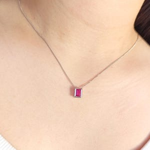 以下が含まれることがあります： 長方形のルビー色のペンダントが付いたシルバーのネックレス。ペンダントはシルバーのフレームにセットされ、繊細なシルバーチェーンから吊り下げられています。白いシャツを着た人がネックレスを着用しています。