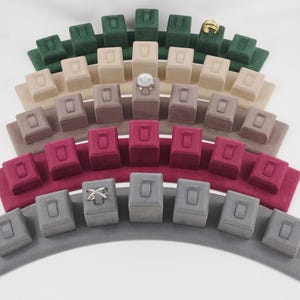 May include: Five curved, tiered jewelry display stands covered in soft velvet, arranged in a fan shape. The colors include dark green, beige, light brown, deep red, and gray. Each stand features multiple individual slots for rings.