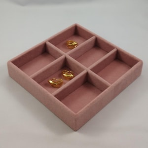 Può includere: Un vassoio portagioie quadrato in velluto rosa polvere con quattro scomparti interni. All'interno del vassoio, tre piccoli orecchini in metallo dorato lucido con un design liscio, arrotondato e a strati riposano in due sezioni.