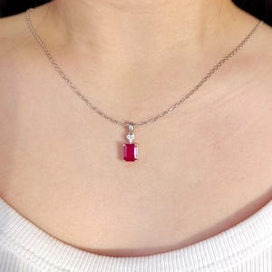 Peut inclure: Un collier en argent avec un pendentif rectangulaire en pierre précieuse rouge foncé. Le pendentif est orné d'une petite pierre transparente au-dessus du rubis. Le collier est porté sur un haut blanc.