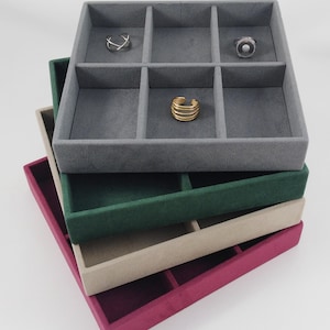 Peut inclure: Plusieurs plateaux à bijoux empilés, de couleurs grise, verte, beige et magenta. Le plateau supérieur contient des bagues. Les plateaux sont divisés en compartiments. Les bagues sont argentées et dorées.