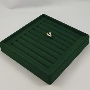 May include: A two-toned ring featuring a silver-colored band that curves upward to meet a small, smooth gold-colored sphere at the top. The ring is displayed on a dark green, rectangular velvet jewelry tray with multiple padded slots.