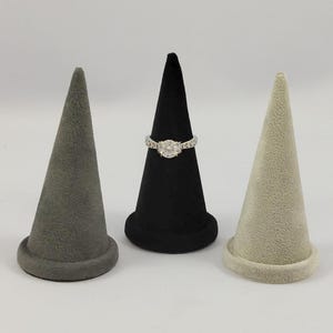 May include: Three cone-shaped jewelry display stands in gray, black, and beige velvet. A silver-toned ring with a round central stone and a decorative band is placed on the black cone. Each stand features a circular base and a tapered top.